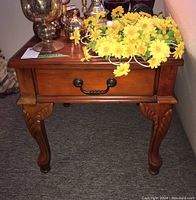 Front view of one wood end table showing faux drawer with black metal handle and carved legs. Yellow artificial flowers on top with other decor items.