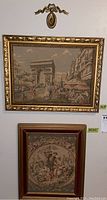 Photo showing both tapestry pieces hung on wall, top with Paris street scene framed in ornate gold frame, bottom pastoral scene framed rectangular with red and gold tones