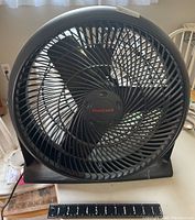 Front view of a black 19 inch Honeywell tilting electric fan with protective grille and blades visible. A measuring tape shows roughly 19 inches diameter.