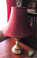 Photo of a single table lamp with a shiny metal base and red patterned fabric shade taken in daylight by a window.