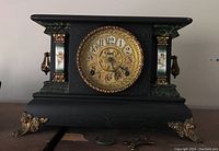 Front view of a vintage black mantle clock showing decorative brass-style feet, gold and green detailed sides, and centered clock face with two winding holes and ornate detailing.