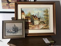 Two framed oil paintings displayed on a table, one larger landscape village scene and one smaller boat scene.