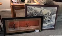 Two framed prints on a couch: a large winter scene of a country store, and a smaller Plakit style print of silhouetted trees on an orange background.