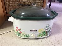 Rival Crock-Pot slow cooker showing white metal base with floral design, control knob, and glass lid on top.