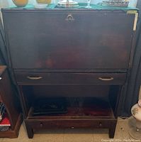 Front view of wooden secretary desk showing fold-down front panel, drawer with brass handles, open shelf and bottom drawer. Visible stains and scratches on surfaces.