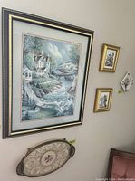 Five wall art pieces hung on a wall with mostly floral and one landscape themed art. Includes a large framed landscape, two smaller floral prints, a ceramic floral piece, and needlepoint with lace under glass in tray frame.
