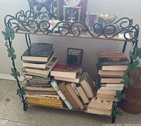 Front view of wrought iron shelf filled with books and decorative items showing floral trim and wicker shelves