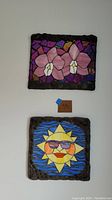 Two concrete mosaic wall art pieces hung vertically. Top piece has pink floral design, bottom piece has sun with sunglasses on blue background.
