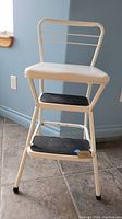 Front angle photo of Cosco step stool showing metal frame, two black steps with non-slip tread, white cushioned seat and backrest