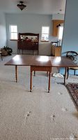 Wooden drop leaf table fully extended in a carpeted room, showing three turned legs and flat surface.