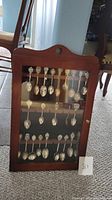 Full view of the wooden hanging spoon display case with glass door showing all spoons inside.