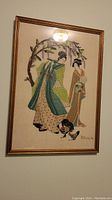 Framed needlework art of two women in kimonos under a flowering tree with a chicken, signed in lower right corner.