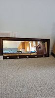 Rectangular solid wood framed mirror with three metal hooks along the bottom front side. Mirror reflecting a kitchen scene. Mirror placed on carpeted floor leaning against a wall.