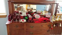 Wide view of Christmas glassware, votives, garland, napkins, and other decorations arranged on wooden dresser with a mirror behind.