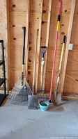 Photo of various long handled garden tools, including rakes, shovels, pitchfork, and blue gardening pot on concrete floor in wooden shed.
