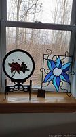 View of two stained glass panels displayed on a windowsill, one circular with red rose design on black stand, and one square with blue flower design in a decorative wire frame.