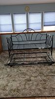 Metal trundle bed frame with black curved decorative headboard and footboard, standing on carpeted floor in front of window. Lower trundle bed frame separated and pushed forward.