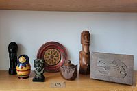 Wood carvings collection with various figures, decorative plate, painted doll, and small lidded jar displayed on a wooden shelf.