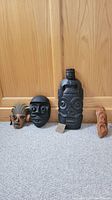 Four African art pieces on carpeted floor against wooden door: three wooden and one metal masks/sculptures.