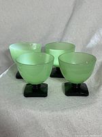Photo showing four vintage jadeite green trophy glasses with black square bases arranged in a row on a light fabric background.