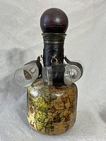 Full view of vintage nautical decanter wrapped in molded Italian leather with antique map printing, topped with large wooden stopper and four small shot glasses hanging from leather loops.