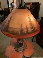 Front view of lamp lit showing reverse-painted glass shade with landscape scene and bronze-tone base
