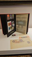 Open black stamp album with vintage Great Britain stamps visible on two pages, some loose stamps and postcards on display