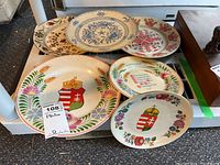 Six decorative plates arranged on a table showing colorful Hungarian folk art motifs and heraldic crests.