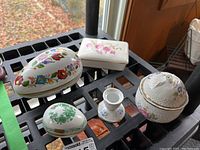 Overview of all four trinket boxes and the candle holder arranged on a black grid surface by a window, showing variety in shapes and colorful hand-painted floral and butterfly designs.