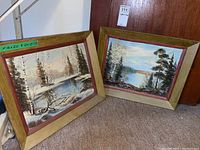 Two framed winter landscape paintings leaning against door on floor