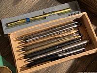 Wood box containing nine metal ballpoint pens, mostly silver, some gold-tone, including black pen. Boxed gold Cross refill above.