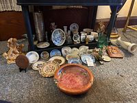 Wide view of the lot showing assortment of plates, figurines, vases, and trinket dishes on carpeted floor under a table.