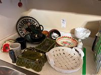 Multiple pottery items on countertop including green planters, black and multicolor decorative plates, white ceramic basket and compote, mini crocks.