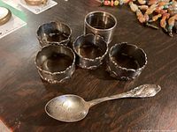 Photo showing five silver napkin rings and one detailed sterling spoon with engraved script and embossed corn handle design.