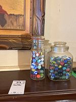 Two glass jars filled with colorful marbles, displayed on a wooden surface near a framed painting.