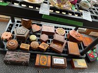 Wide view of 17 assorted carved wood and other keepsake boxes on black grate shelf showing shapes, sizes, and surface designs.
