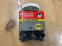 Plastic jar with white lid containing 300 black GB coaxial staples for wood with clear labeling and quantity count