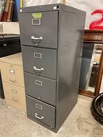 Front and angled view of a tall gray metal file cabinet showing four drawers with handles and label holders. Cabinet shows some wear and scratches.