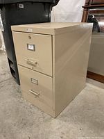Front and side view of small beige two-drawer metal file cabinet next to a black trash bin and wood frame mirror.