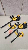 Four Dewalt work clamps displayed on a flat surface, showing two sizes including two large and two smaller clamps with yellow and black color scheme and Dewalt branding.