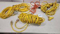 Four heavy duty extension cords arranged on table showing cords coiled, three in yellow and one orange, with visible plugs and outlet ends.
