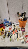Full overview of the painting supplies lot on a table including paint brushes, foam applicators, paint tape, paintbrush cleaner, and scrapers.