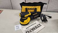 Dewalt orbit palm sander with cord, carrying bag, and manual on table.
