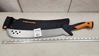Fiskars machete fully sheathed, showing the branded sheath and ergonomic handle with ruler for size reference.