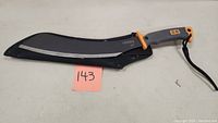Photo showing full machete with curved black sheath, gray blade edge visible, handle has orange and gray accents with BG logo.