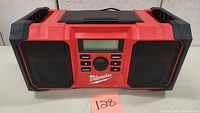 Front view of Milwaukee jobsite clock radio showing red and black design, speakers, digital display, and control buttons.