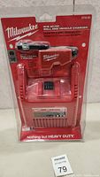 Front view of Milwaukee M18 AC/DC charger in sealed packaging showing charger and accessory plugs.