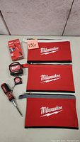 Overview of all items in the lot showing three Milwaukee red tool bags, two tape measures, one screwdriver, and one unopened screwdriver bit set.