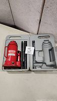 Red Motomaster 12 ton bottle jack inside gray molded plastic carrying case with detachable handle sections.