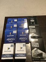 Lot of new Pencil Buddies pencils in original boxes on a table, including blue boxes of kids triangular pencils, white boxes of golf pencils, and black boxes of sketching pencils.